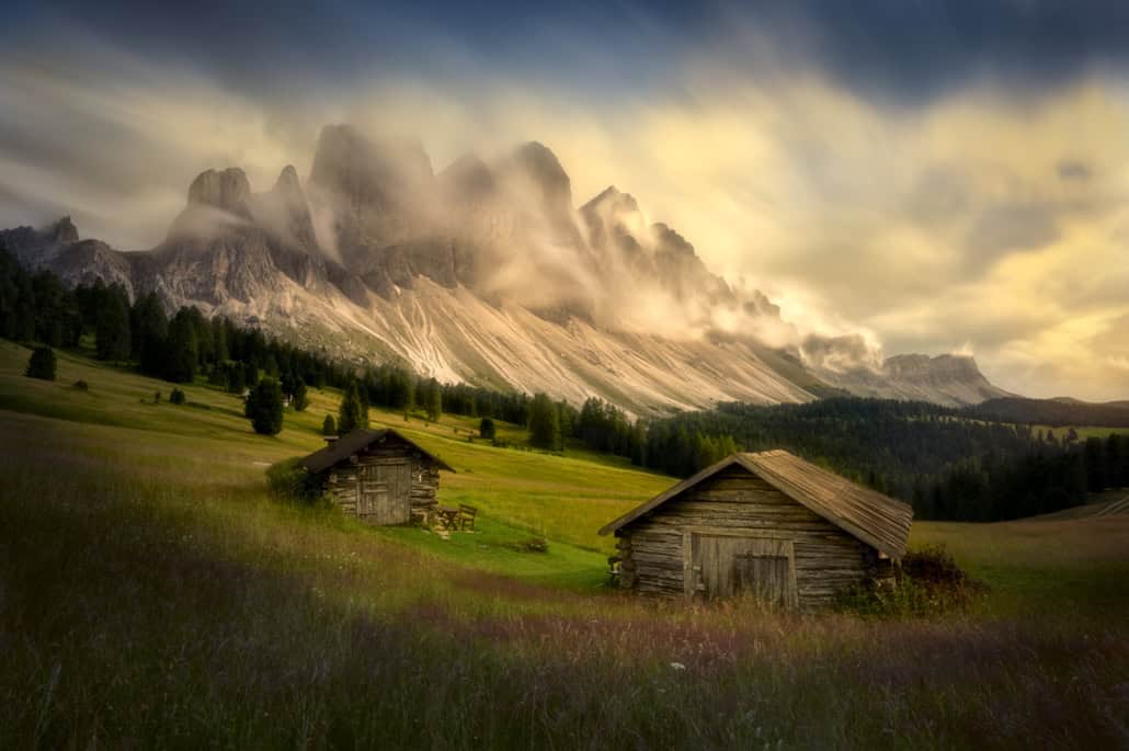 Barevné horské panorama s dřevěnými chatkami, mlhou a rozmazaným nebem v pozadí. Letní Dolomity - fotokurzy a fotografické workshopy